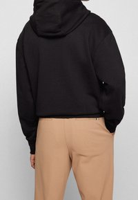 Persona con un hoodie nero e pantaloni beige, in piedi con una mano in tasca, vista di spalle su uno sfondo chiaro e uniforme.