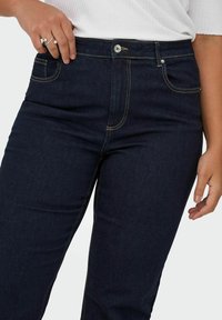 Tumši zilie džinsi ar augstu jostasvietu, kam ir dzeltena šūšana, priekšējās kabatas un metāla pogas aizdare. Gluda denima tekstūra.