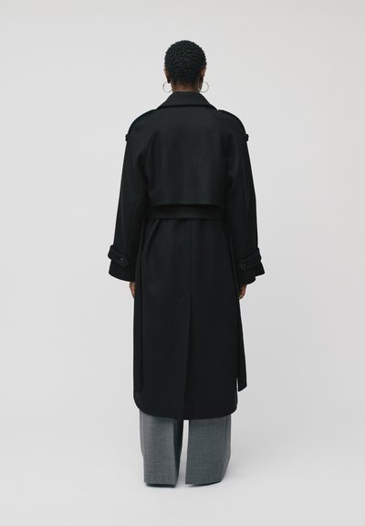 Trench coat negru cu un design fluid, mâneci largi și talie accesorizată cu un brâu. Materialul pare texturat; expus din spate împreună cu pantaloni.