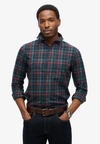 Geruit overhemd in groen, blauw en bordeauxrood met lange mouwen, een button-down kraag en een borstzak; gecombineerd met donker denim en een bruine riem.