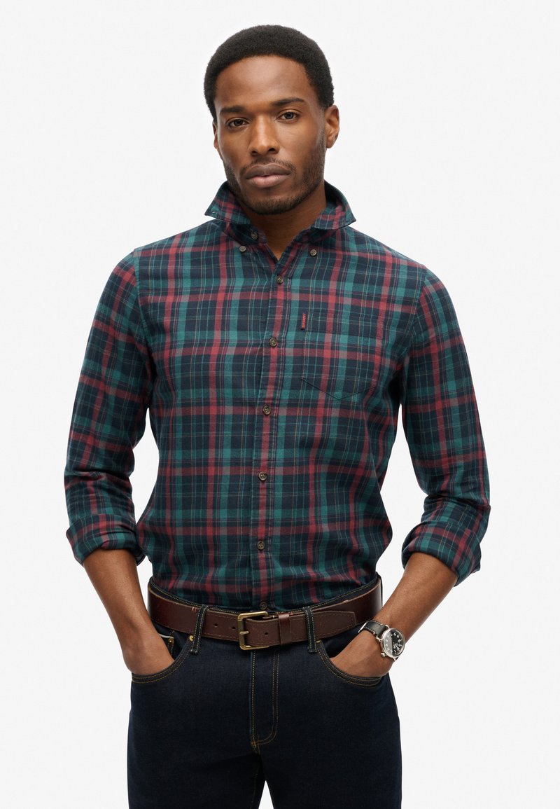 Geruit overhemd in groen, blauw en bordeauxrood met lange mouwen, een button-down kraag en een borstzak; gecombineerd met donker denim en een bruine riem.