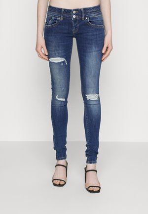 Skinny blue denim jeans with distressed details on the knees, featuring a mid-rise waist and two buttons at the front, paired with black heels.