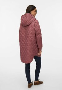 Ženska, oblečena v dolgo, prešito vijolično jakno s kapuco, temne oprijete jeans hlače in rjave usnjene čipkaste čevlje, stoji pred belim ozadjem.