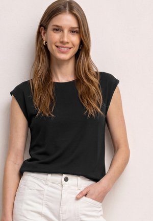 Jeune femme aux longs cheveux châtain clair sourit, portant un haut noir sans manches et un pantalon blanc, debout contre un mur clair uni.