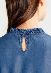 Blouse en denim avec un col à volants, un dos ajouré et une texture douce dans une teinte bleu moyen.