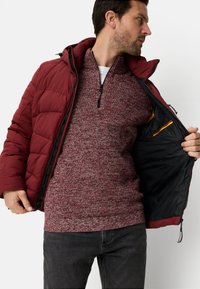 Bordowa kurtka puchowa z kapturem na bordowym swetrze z dzianiny. Sweter ma teksturowany wzór; wnętrze kurtki jest czarne z pomarańczowym akcentem.