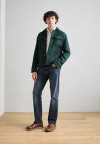 Giacca verde con texture, due tasche sul petto, chiusura con bottoni, abbinata a jeans scuri e scarpe marroni, in piedi su un pavimento di legno.