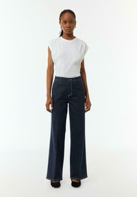 Witte mouwloze top gecombineerd met donkerblauwe wijde jeans, met subtiele stiksels en een hoge taille, aangevuld met zwarte instapschoenen.