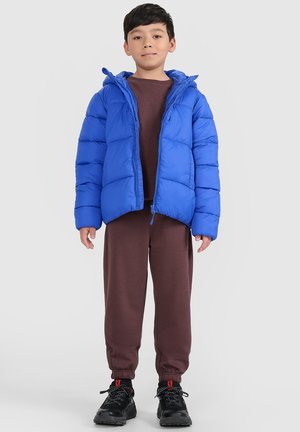 Dziecko stoi przodem, ubrane w jaskrawo niebieską kurtkę puchową na brązowym dresie i czarnych sneakersach, na jednolitym tle.