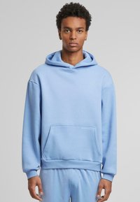 Felpa blu chiaro realizzata in morbido tessuto, con tasca frontale e cappuccio regolabile. Il design include un dettaglio testuale discreto sulla tasca.
