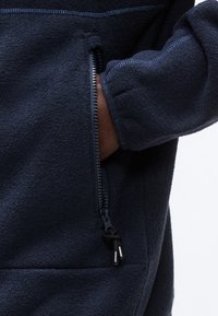 Μανίκι από σκούρο μπλε fleece ζακέτα με ένα χέρι μερικώς μέσα σε κάθετη τσέπη με φερμουάρ, με μαύρα κορδόνια φερμουάρ.