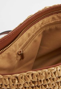 Bolso de paja tejido con un forro de tela marrón; presenta un bolsillo con cremallera y herrajes en tono dorado. El interior es suave y de color tostado.