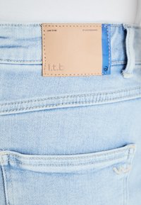 Light blue denim jeans featuring a tan leather patch with embossed text and a blue accent at the waistband. Distinct stitching and back pocket detail.
