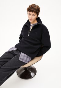Pullover in pile nero con colletto a zip e un logo discreto. Indossato sopra una camicia grigia a scacchi, abbinato a jeans scuri, su una sedia curva.