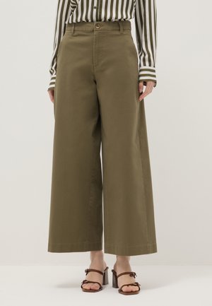 Vrouw draagt olijfgroene wijde broek met hoge taille, gecombineerd met een gestreept overhemd en bruine sandalen met hak.