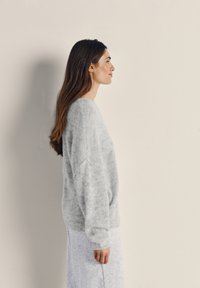 Woman with long brown hair wearing a soft gray sweater and skirt, standing sideways against a plain beige background.