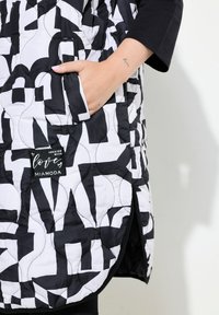 Quiltad ytterklädsel i svart och vitt med ett djärvt grafiskt mönster. Har en sidficka och en etikett med texten "Skapat med kärlek, Miamoda."