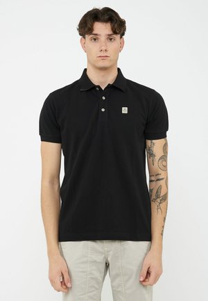 Giovane uomo con capelli corti e scuri che indossa una polo nera e pantaloni chiari, mostrando i tatuaggi sull'avambraccio destro.