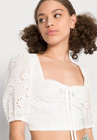 Top branco cropped com mangas bufantes, bordado em ilhós, decote quadrado, detalhe com cordão na frente, tecido leve.