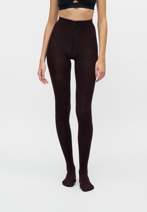 Femme portant des collants opaques marron foncé à taille haute et un crop top noir, debout devant un fond clair uni.