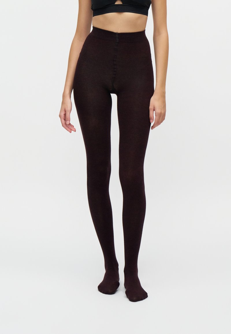 Femme portant des collants opaques marron foncé à taille haute et un crop top noir, debout devant un fond clair uni.