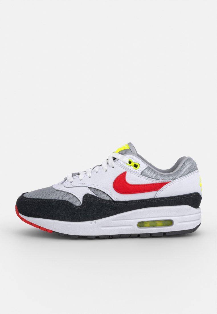 Les baskets Nike Air Max 1 présentent une tige en mesh gris, des accents en suède noir, du cuir blanc, un logo rouge et des détails jaunes, avec un coussin d'air visible.