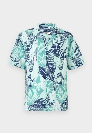 Muž s krátkými blond vlasy, který nosí modro-zelenou košili s krátkým rukávem a potiskem listů, černé šortky po kolena, bílé ponožky a béžové tenisky.