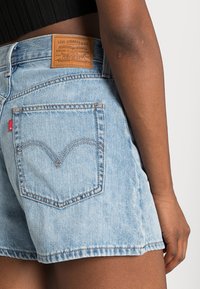 Pantalones cortos de mezclilla azul claro con cintura alta, que cuentan con un parche de marca de cuero y costura distintiva en el bolsillo trasero. Detalle de dobladillo deshilachado visible.