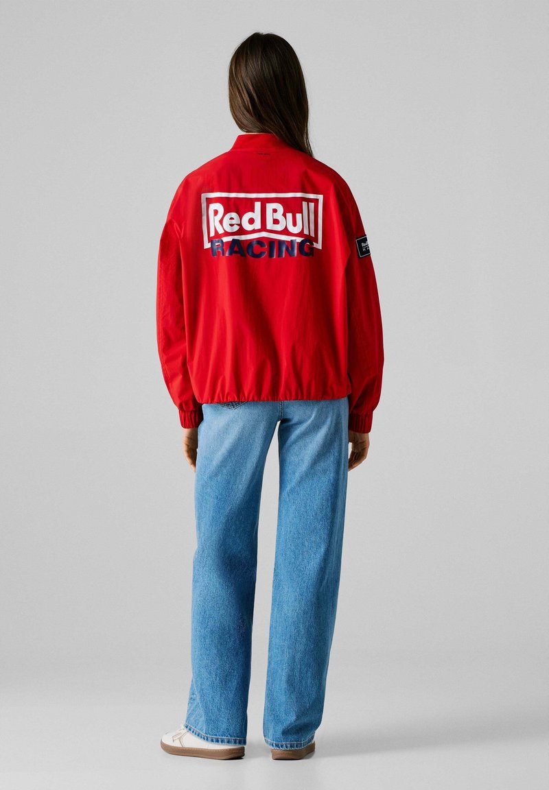 Veste bomber rouge en tissu rouge vif avec le logo "Red Bull RACING" dans le dos. Associée à un jean bleu décontracté et des chaussures blanches.