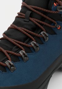 Nærbilde av blå tursko med svarte og oransje mønstrede lisser trådet gjennom metallkroker og øyeknopper.