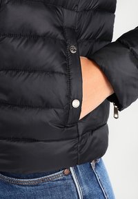 Svart puffjacka med kviltad textur, synlig knapp och dragkedja ficka, bärs över ljus denimjeans. Minimalistisk design.