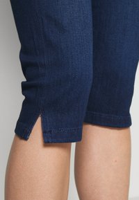 Tummansiniset denim-caprit, joissa on istuva muotoilu, pienen sivuv splitting reunassa ja hienovaraiset tikkaus yksityiskohdat. Sujuva pinta.