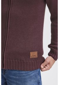 Maglione lavorato a maglia color bordeaux con polsini e orlo a coste. Presenta una patch in pelle sul lato inferiore. Il tessuto ha una finitura testurizzata.