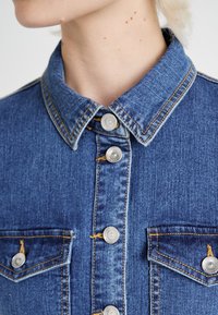 Τζάκετ από τζιν με αιχμηρό γιακά, ασημένιες κουμπωτές ταινίες και αντίθετ�η κίτρινη ραφή. Διαθέτει δύο τσέπες στο στήθος και κλασικό μπλε πλύσιμο.