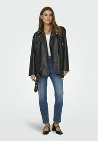 Zwarte leren motorjack met brede revers en zilveren ritsdetails, gedragen over een wit shirt en gecombineerd met blauwe spijkerbroeken en zwarte schoenen.