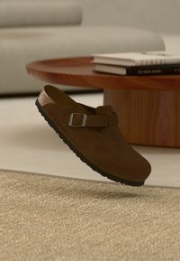 Καφέ σουέντ clog χωρίς κορδόνια με μαύρη σόλα και ασημένια αγκράφα, αιωρούμενο πάνω από μπεζ υφασμάτινο χαλί, ξύλινο τραπεζάκι καφέ και ανοιχτό γκρι καναπέ στο βάθος.