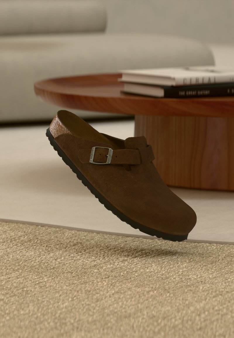 Καφέ σουέντ clog χωρίς κορδόνια με μαύρη σόλα και ασημένια αγκράφα, αιωρούμενο πάνω από μπεζ υφασμάτινο χαλί, ξύλινο τραπεζάκι καφέ και ανοιχτό γκρι καναπέ στο βάθος.