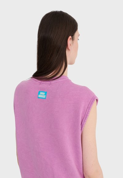 Femme aux longs cheveux bruns portant un haut lavande sans manches avec une petite étiquette bleue sur le haut du dos, sur un fond gris uni.