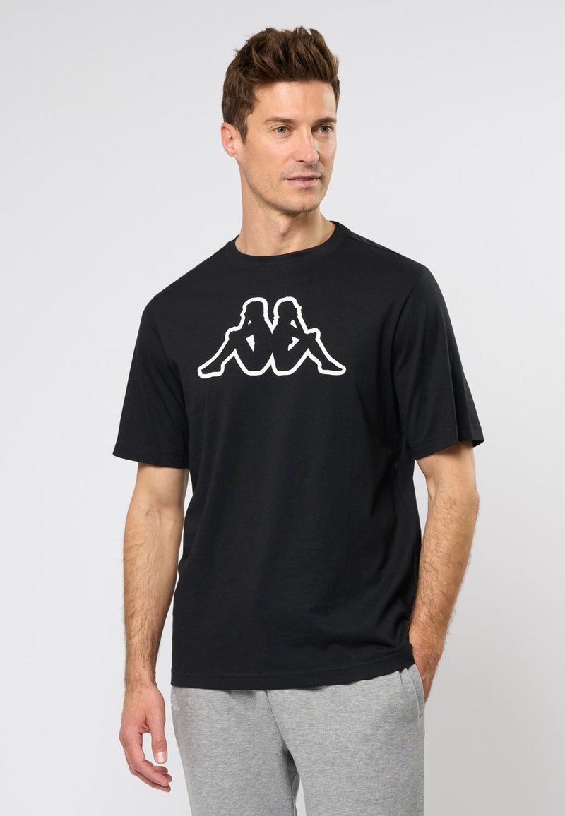 Zwarte katoenen t-shirt met een groot wit grafisch logo van twee zittende figuren. Ronde hals, korte mouwen, relaxed fit. Grijze sweatpants zichtbaar.