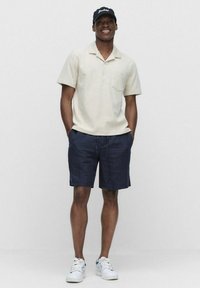 Jasno beżowa koszula z krótkim rękawem, z kołnierzykiem i kieszenią na piersi, zestawiona z granatowymi szortami oraz białymi trampkami z zielonymi akcentami.
