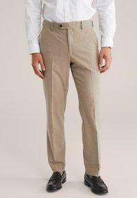 Homme portant un pantalon beige sur mesure, une chemise blanche à boutons et des mocassins en cuir marron foncé, se tenant sur un fond neutre.