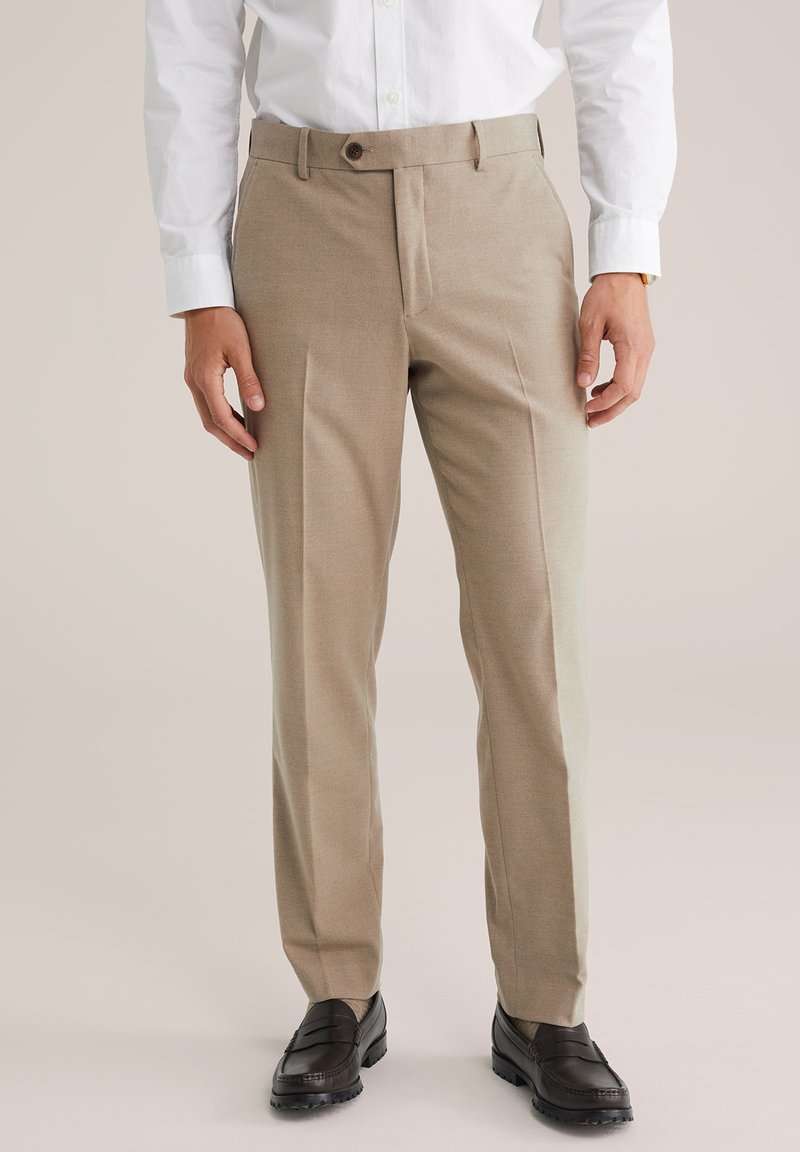 Homme portant un pantalon beige sur mesure, une chemise blanche à boutons et des mocassins en cuir marron foncé, se tenant sur un fond neutre.