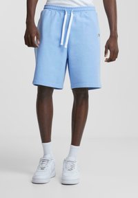 Shorts en mélange de coton bleu clair avec une taille élastique, des cordons de serrage blancs et des poches latérales, comportant un petit logo sur la jambe gauche.