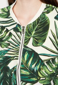 Close-up van een jack met rits aan de voorkant, bedrukt met grote groene tropische bladeren op een witte stof, gedragen op de bovenste borst.
