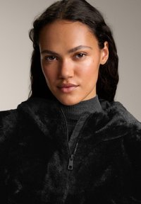 Hanorac din blană sintetică neagră cu închidere cu fermoar, având o textură moale și un guler înalt, purtat peste un top gri cu textură în valuri. O notă de strălucire pe material.