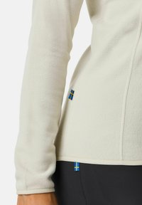 Fleece jacket in off-white with a soft texture, featuring blue-yellow Swedish flag patches at the hem and side. Contrast stitching detail.