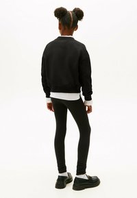 Niña con dos coletas esponjosas, vestida con un sudadera negra sobre una camiseta blanca, leggins negros, calcetines blancos y mocasines negros, de pie de espaldas.
