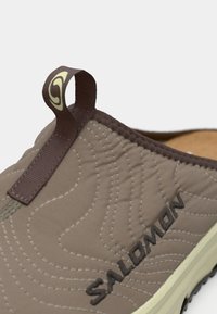 Brune slip-on sko med quiltet design, teksturert stoff, fremtredende sømmer og en løfreetikett med logo. Sålen har gripe detaljer.