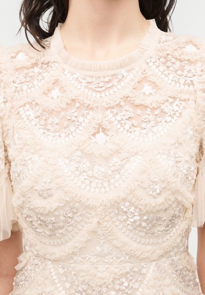 Διαφανές μπεζ μπλουζάκι από δαντέλα με floral σχέδια, βολάν και παγιέτες, που δείχνει το λαιμό και το πάνω μέρος του κορμού.