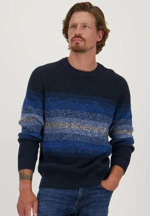 Pull en tricot dégradé de bleu navy, bleu et gris ; présente un col rond et des poignets côtelés ; tissu texturé. Porté avec un jean en denim.
